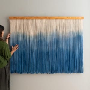 Puede incluir: Un gran tapiz de pared con una parte superior de madera y largos hilos verticales. El hilo pasa de blanco en la parte superior a un azul profundo en la parte inferior, creando un efecto degradado. La pieza está montada en una pared gris claro.