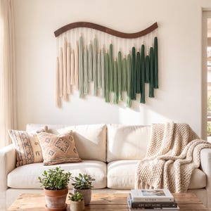 May include: A wall hanging featuring a curved dark wood top bar with hanging cotton cords in an ombre color gradient, transitioning from light cream to sage green and deep forest green, accented with small wooden beads on each cord strand.
