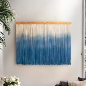 May include: A large wall hanging with a natural wood top and a gradient design. The top portion is off-white, transitioning to a deep blue at the bottom. The hanging is made of many vertical strands.