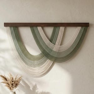 May include: A wall hanging featuring a dark brown wooden bar with strands of green and off-white yarn cascading downwards. The yarn is arranged in a curved, layered design, creating a modern, textured aesthetic. The wall decor is a neutral color palette.