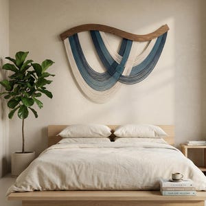 May include: A modern bedroom scene featuring a wooden bed frame with cream-colored bedding. Above the bed, a wall hanging with a wooden top and blue and white woven strands adds a decorative touch. A potted plant and books complete the minimalist aesthetic.