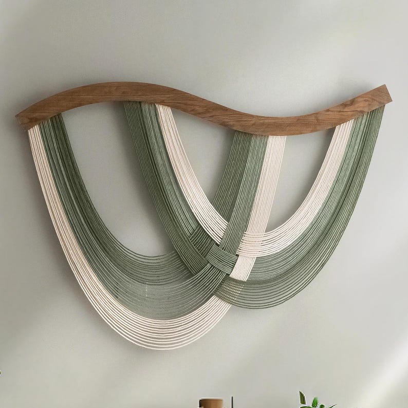 May include: A woven wall hanging featuring a curved wooden top and cascading strands in shades of green and off-white. The strands are intricately woven together, creating a unique and textured design. This piece adds a touch of modern art to any space.
