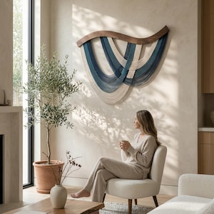 May include: A modern wall hanging with a wooden frame and woven blue and cream-colored strands. The artwork is displayed in a living room with a neutral color palette, a small tree, and a woman sitting in a chair.