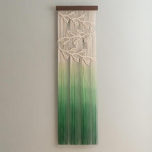 May include: A macrame wall hanging with a wooden top. The design features cream-colored leaves and a gradient of green threads. The threads transition from light green to a darker shade, creating a visually appealing ombre effect. This piece is a decorative wall art.