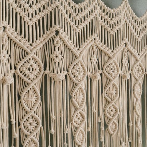 May include: A beige macrame wall hanging with a geometric pattern. The hanging is made of thick cotton cord and has a fringe at the bottom.