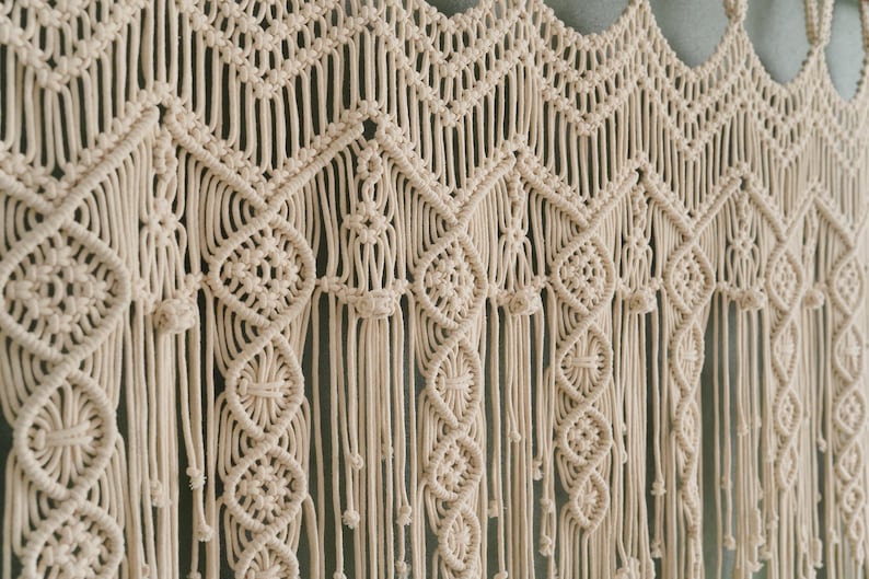 May include: A beige macrame wall hanging with a geometric pattern. The hanging is made of thick cotton cord and has a fringe at the bottom.