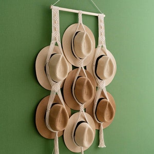 May include: A macrame hat rack with nine straw hats. The hats are in shades of tan and brown, with black bands. The rack is made of natural-colored macrame and hangs against a green wall.