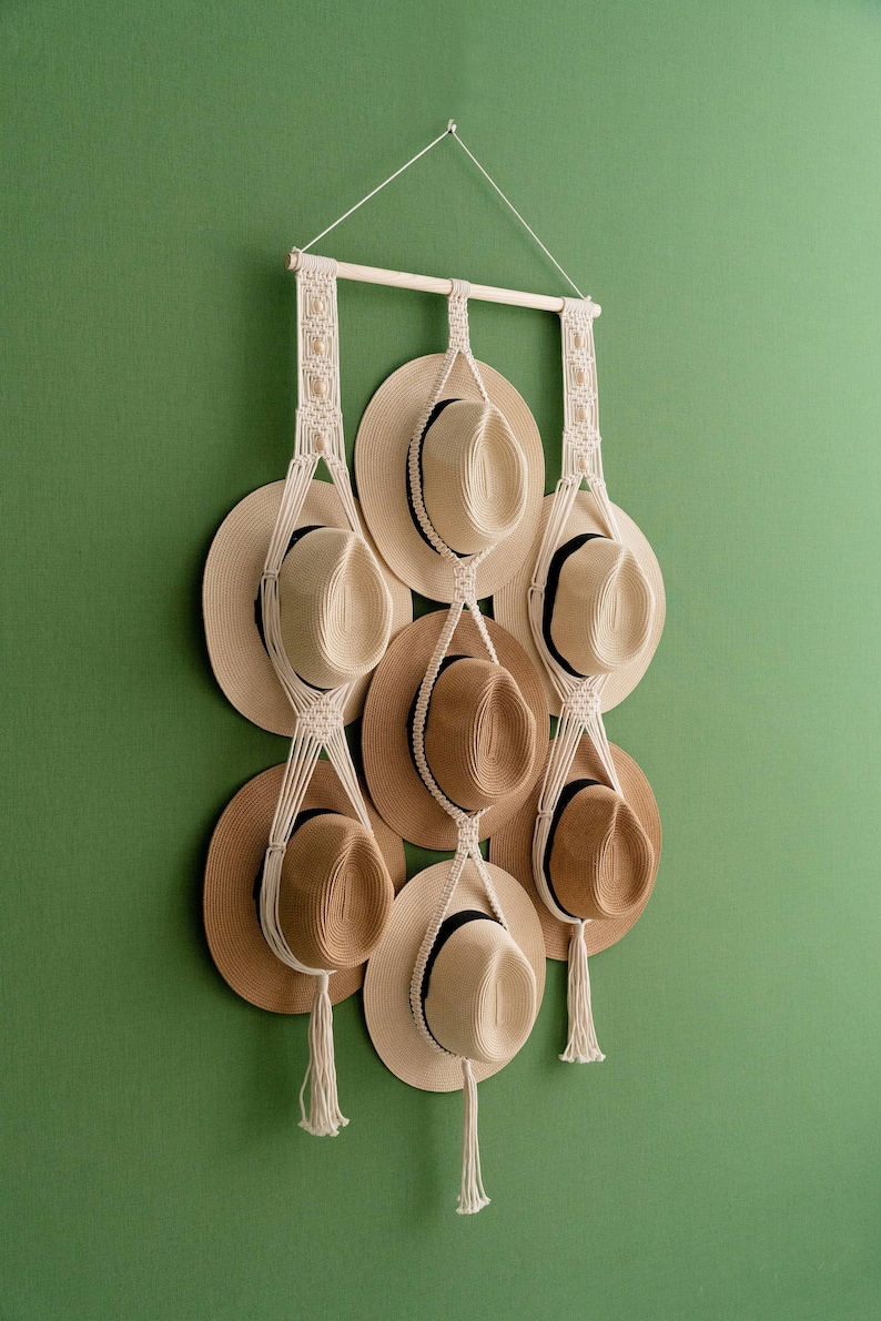 May include: A macrame hat rack with nine straw hats. The hats are in shades of tan and brown, with black bands. The rack is made of natural-colored macrame and hangs against a green wall.