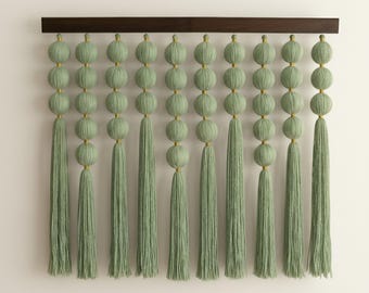 May include: A decorative wall hanging featuring rows of sage green yarn spheres and tassels suspended from a dark brown wooden bar. The spheres are connected with gold-colored accents, creating a textured, artistic design.