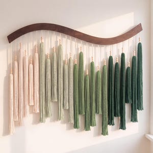 May include: A wall hanging featuring a curved dark wood bar with multiple hanging yarn tassels in a gradient of colors ranging from light cream and soft sage green to deep forest green, accented with small natural wooden beads on each cord.