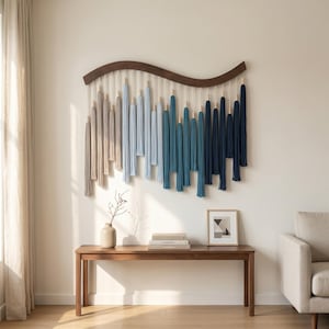 May include: A wall hanging featuring a curved dark wood top bar with hanging cotton cords in an ombre color gradient from beige to light blue, teal, and dark navy blue. Each cord is accented with small wooden beads at varying heights.