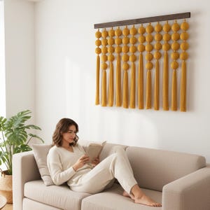 Könnte beinhalten: Ein Wandbehang mit einer horizontalen Leiste aus dunklem Holz, an der mehrere vertikale Stränge aus senfgelbem Garn hängen. Jeder Strang besteht aus einer Reihe runder, texturierter Garnkugeln, die in langen, fließenden Garnquasten enden.