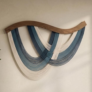 Puede incluir: Un tapiz de pared con una barra superior de madera curvada y teñida de oscuro, de la que cuelgan múltiples hilos de cuerda en tonos crema, azul claro y azul marino oscuro. Las cuerdas están dispuestas en bucles superpuestos en forma de U.