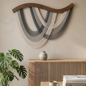 May include: A modern wall hanging featuring a dark wood curved top with cascading strands in shades of gray and white. The artwork is mounted on a neutral-colored wall. The image also shows a wooden cabinet, a plant, and a coffee table with a cup of coffee.