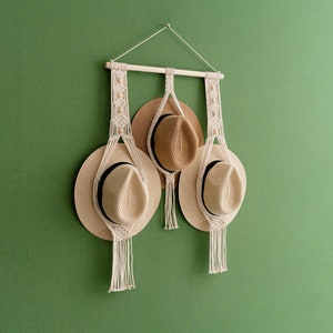 May include: A macrame hat rack with three straw hats. The rack is made of natural-colored rope and a wooden dowel. The hats are beige with black bands. The rack is hanging on a green wall.
