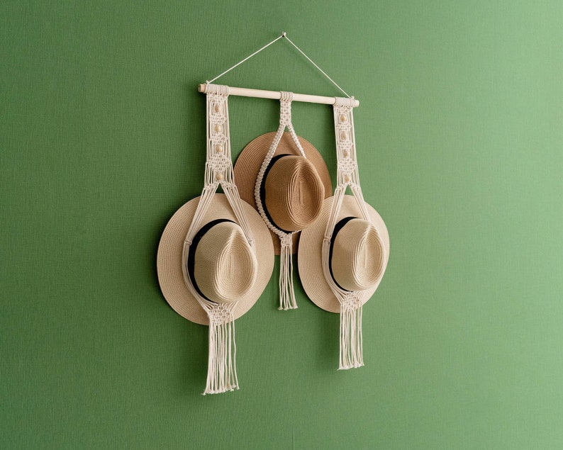 May include: A macrame hat rack with three straw hats. The rack is made of natural-colored rope and a wooden dowel. The hats are beige with black bands. The rack is hanging on a green wall.