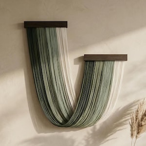 Könnte beinhalten: Ein Wandbehang mit zwei horizontalen Leisten aus dunklem Holz, an denen lange, vertikale Baumwollkordeln hängen. Die Kordeln verlaufen farblich von tiefem Waldgrün über Salbeigrün bis hin zu Cremeweiß und bilden eine geschwungene U-Form.