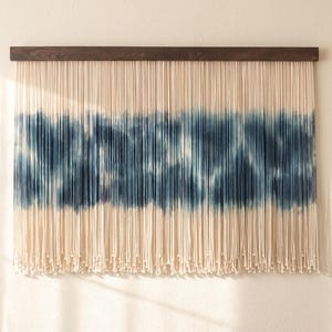 May include: A wall hanging featuring a horizontal dark brown wooden bar from which numerous off-white cotton cords are suspended. A wide, horizontal band of indigo blue tie-dye pattern is visible across the center of the hanging cords.