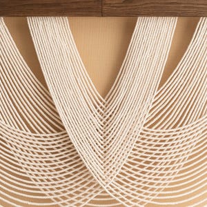 May include: Cream-colored macrame wall hanging with a geometric design. The piece features numerous vertical strands of rope that curve and fan out, creating a textured, decorative element. A dark wood element is at the top.