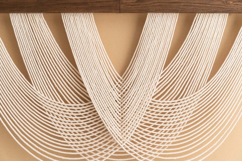 May include: Cream-colored macrame wall hanging with a geometric design. The piece features numerous vertical strands of rope that curve and fan out, creating a textured, decorative element. A dark wood element is at the top.