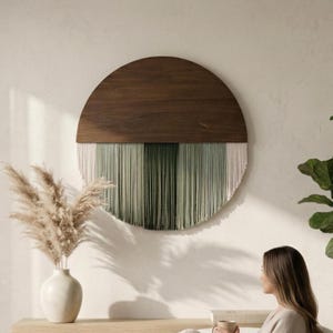 May include: A circular wall hanging featuring a dark brown wooden semi-circle top and a bottom section composed of hanging cotton cords in gradient shades of cream, sage green, and dark forest green, creating a layered fiber art piece.