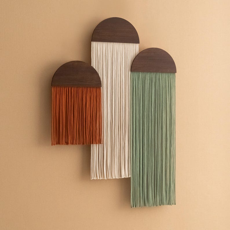 May include: Three wall hangings with wooden half-circle tops and long strands of yarn in orange, white, and green.