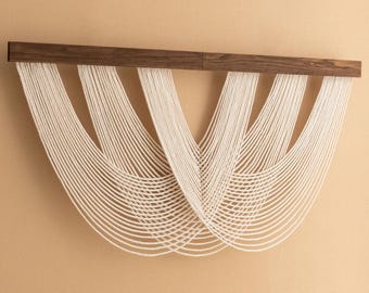 May include: A white macrame wall hanging with a curved design, suspended from a dark wood bar.