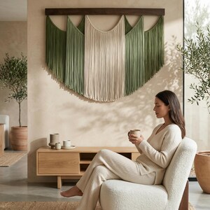 May include: A large macrame wall hanging with a dark brown wooden bar. The macrame features a central panel of cream-colored fringe flanked by panels of green fringe. A woman sits in a white chair.
