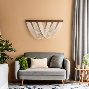 May include: A modern living room scene featuring a gray sofa with decorative pillows and a green throw. A woven wall hanging with a dark wood top and white strands hangs above the sofa. A small wooden side table and a potted plant are also visible.