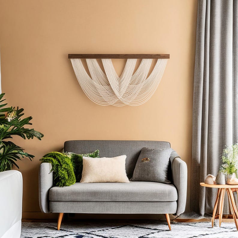 May include: A modern living room scene featuring a gray sofa with decorative pillows and a green throw. A woven wall hanging with a dark wood top and white strands hangs above the sofa. A small wooden side table and a potted plant are also visible.