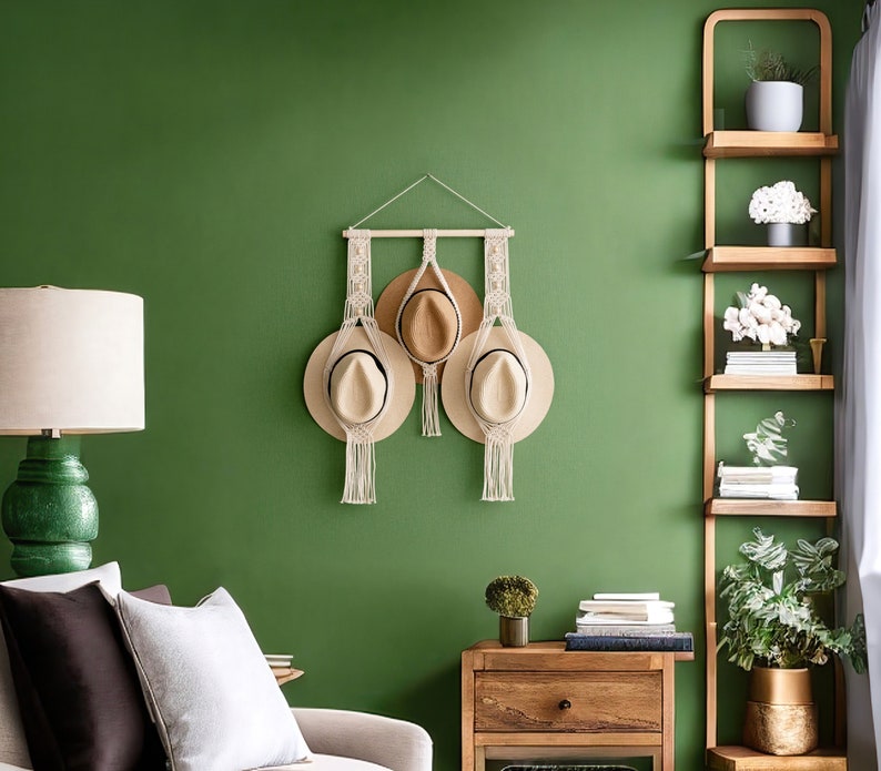 May include: A macrame hat holder with three hats hanging on a green wall. The hats are beige and brown, and the macrame is cream-colored. A wooden shelf with plants and books is to the right. A green lamp and a side table are in the foreground.
