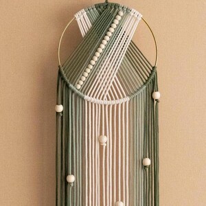 May include: A macrame wall hanging with a circular wooden frame and white and green yarn. The hanging features a geometric pattern and is decorated with small wooden beads.