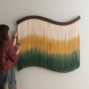 May include: A large, wall-mounted textile art piece with a wavy, dark brown wooden frame. The artwork features vertical strands of cream, yellow, and green fibers, creating a gradient effect. The piece is being held by a person.