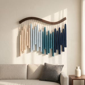 May include: A wall hanging featuring a curved dark wood bar with hanging cotton cords in an ombre color gradient. The cords transition from cream to light blue, teal, and dark navy blue, accented with small wooden beads at various heights.