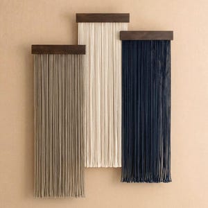 May include: Three modern wall hangings. Each has a dark brown wooden top with long, vertical strands. The strands are in beige, navy blue, and olive green. The wall hangings would suit a contemporary home.
