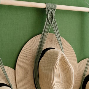 May include: A macrame hat rack with three hanging sections. The rack is made of a light-colored wooden dowel and sage green macrame cord. Three beige sun hats with black bands are displayed on the rack against a green wall.