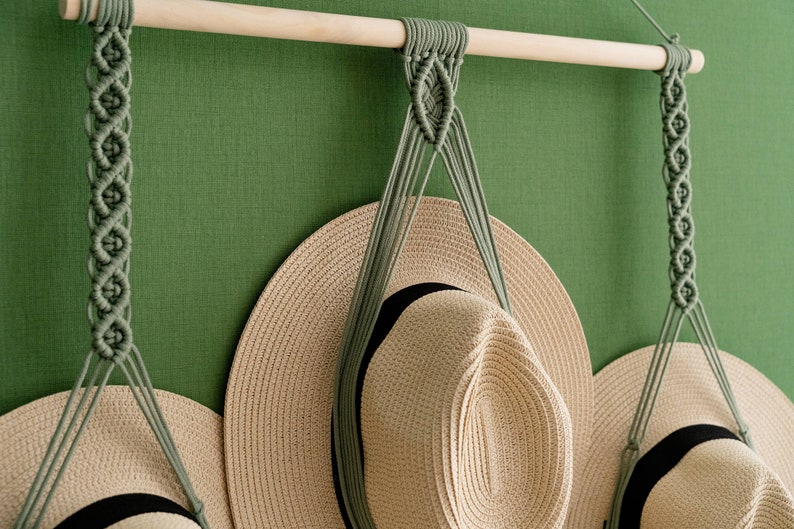 May include: A macrame hat rack with three hanging sections. The rack is made of a light-colored wooden dowel and sage green macrame cord. Three beige sun hats with black bands are displayed on the rack against a green wall.