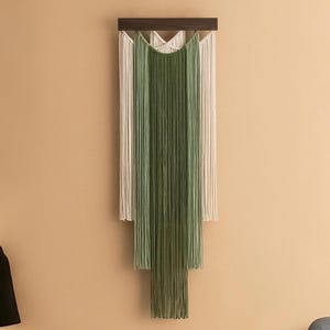 May include: A wall hanging made of woven yarn in shades of green and white. The yarn is arranged in a cascading pattern, creating a modern and minimalist design.