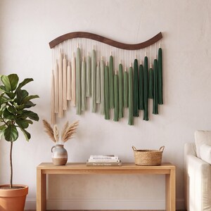 May include: A wall hanging featuring a curved dark wood bar with hanging fiber tassels in an ombre color gradient ranging from cream to sage green and deep forest green, accented with small wooden beads on each individual hanging cord.