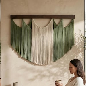 May include: A wall hanging featuring long, vertical strands of fiber suspended from a dark brown horizontal wooden bar. The design consists of alternating sections of sage green and cream-colored cord, arranged in a layered, scalloped pattern.