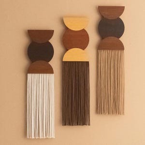 Op de afbeelding: Drie abstracte wandkleden met een modern design uit het midden van de eeuw. Elk stuk heeft een combinatie van houten halve cirkels en cirkels in bruin, zwart en tan, met verticale franjes in wit, bruin en beige. De achtergrond is een neutraal beige.