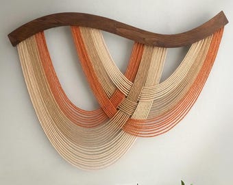 May include: A modern wall hanging featuring a curved, dark wood frame. Strands of orange and beige yarn are woven and draped, creating a textured, abstract design. The artwork is mounted on a light-colored wall, adding a touch of contemporary art to the space.