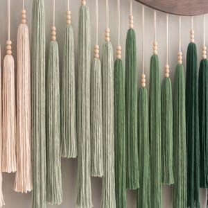 May include: A wall hanging featuring a series of long, vertical yarn tassels in a gradient of colors ranging from pale blush pink to sage green and deep forest green. Each tassel is suspended by a cord accented with three small wooden beads.