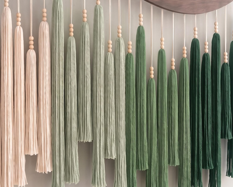 May include: A wall hanging featuring a series of long, vertical yarn tassels in a gradient of colors ranging from pale blush pink to sage green and deep forest green. Each tassel is suspended by a cord accented with three small wooden beads.