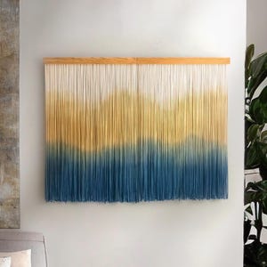 Könnte beinhalten: Ein dekorativer Wandbehang mit Farbverlauf. Der obere Teil ist weiß, geht in Gelb über und endet unten in Blau. Die Fäden sind an einem Naturholzrahmen befestigt und bilden ein strukturiertes Kunstwerk.