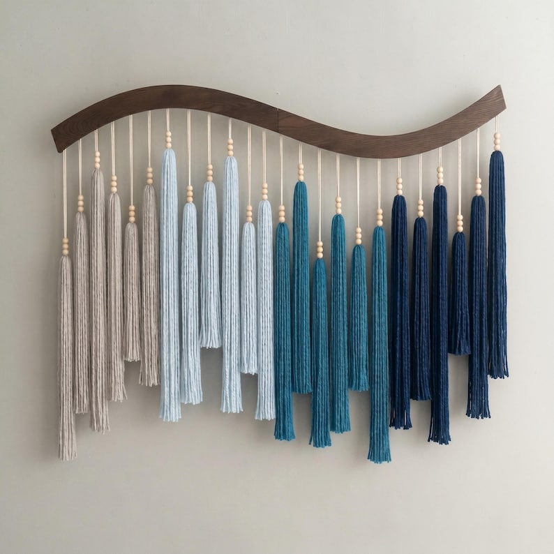 May include: A wall hanging featuring a curved dark wood bar with multiple hanging yarn tassels in an ombre color gradient. The tassels transition from light beige to light blue, teal, and deep navy blue, each accented with small wooden beads.
