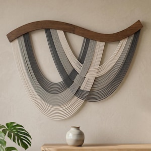 Pode incluir: Uma tapeçaria de parede decorativa com uma parte superior de madeira castanha escura curva. O design apresenta fios entrelaçados de cordões brancos, cinzentos e cinza escuro. A obra de arte é exibida contra uma parede de tom neutro.
