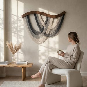 May include: A modern living room scene featuring a large, abstract wall hanging with a dark wood frame and woven strands in shades of gray and cream. A woman sits in a cream-colored chair, holding a mug. A wooden side table and rug complete the space.