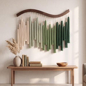 May include: A wall hanging featuring a curved dark wood bar with hanging cotton cords in an ombre color gradient from cream to light green and dark forest green, accented with small wooden beads at the top of each tassel strand.