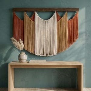 May include: A large macrame wall hanging with a wooden bar, featuring a color palette of rust, tan, and cream. Below, a light wood console table with a vase and a book titled "The Art of Slow Living".